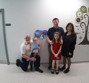 Little boy holding certificate with family standing next to bell