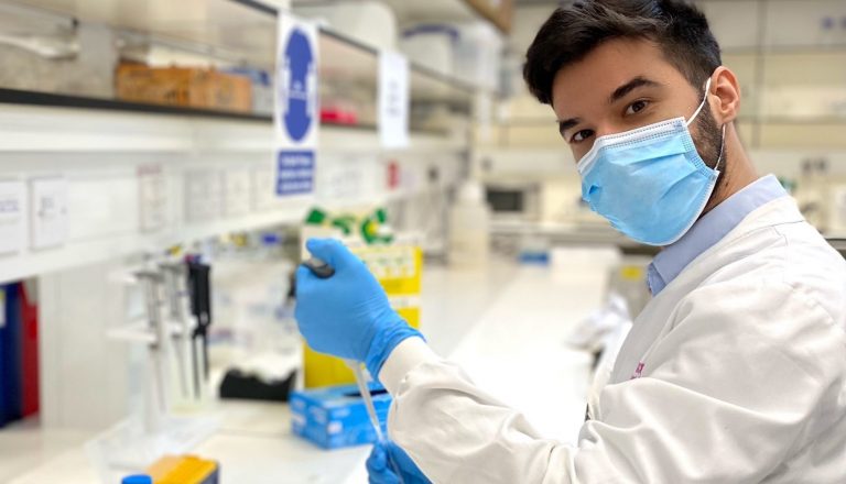 Researcher wearing mask and white lab coat holding pipette