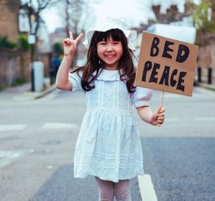 Astrid smiling wearing white hat standing in the road with a peace v sign and holding a bed peace poster min